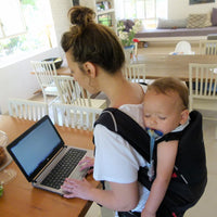 A baby sleeping in a Black Cococho baby carrier while baby is carried on his moms back while mom is working using her laptop.