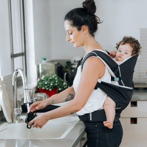 A mom is back carrying her baby girl in the Cococho Baby Carrier and washing the dishes 