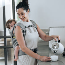 Load image into Gallery viewer, Mom is carrying her baby on the back with the Cococho baby carrier and making a cup of coffee for herself