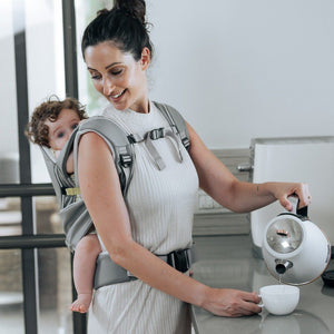 Mom is carrying her baby on the back with the Cococho baby carrier and making a cup of coffee for herself